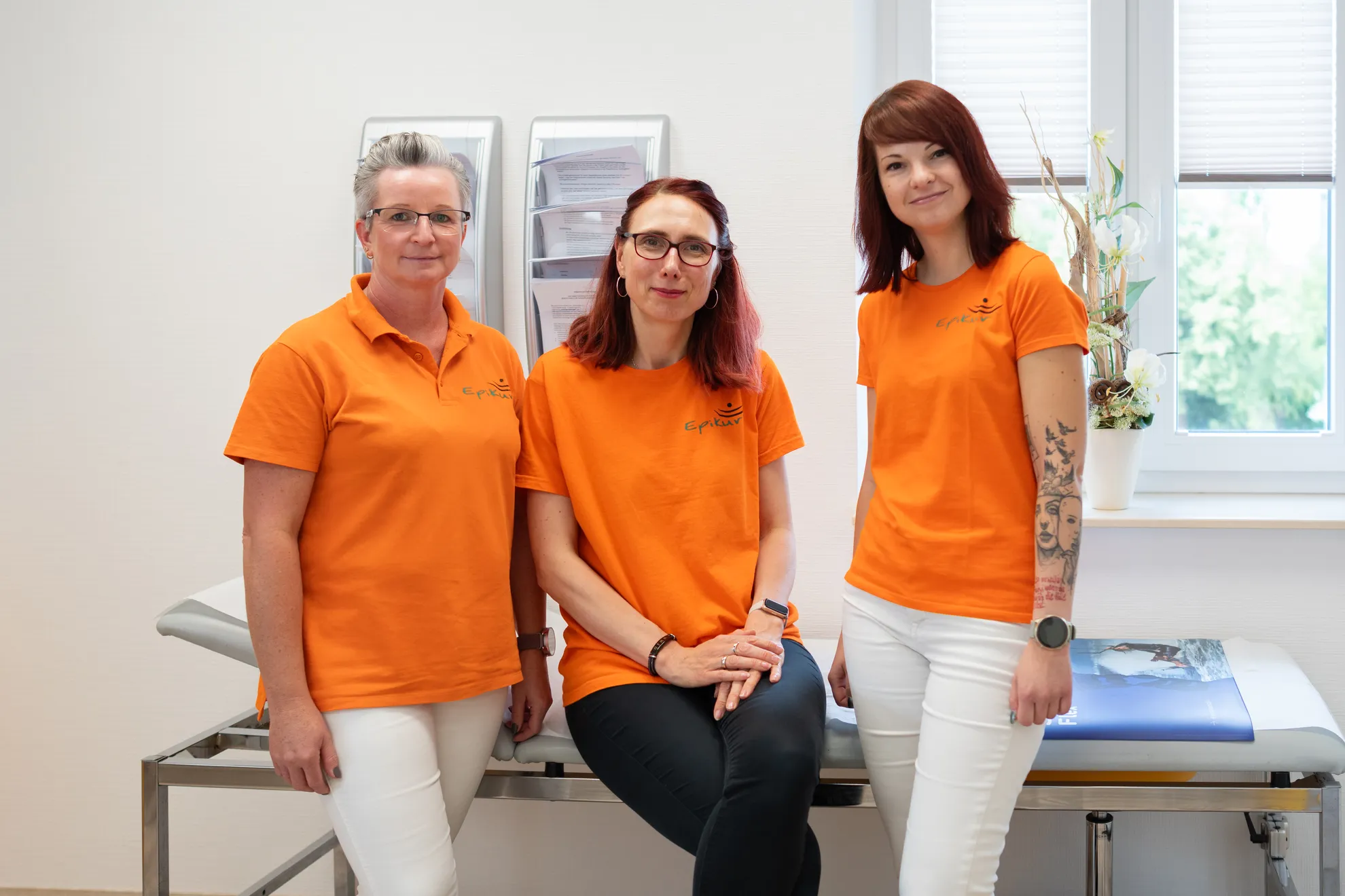 3 Personen anlehnend auf einer Liege im Behandlungsraum lächelnd in die Kamera